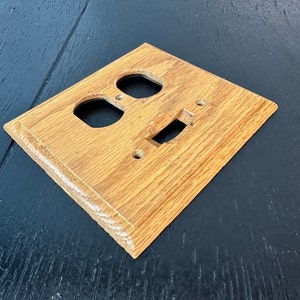 May include: A wooden electrical outlet cover with two circular openings for outlets and a rectangular opening for a light switch. The cover is made of light brown wood with visible grain.