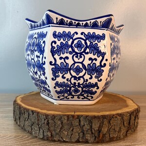 Puede incluir: Una maceta de cer&aacute;mica blanca y azul con borde festoneado. La maceta presenta un patr&oacute;n floral azul repetido. La maceta est&aacute; sobre una rodaja de madera redonda.