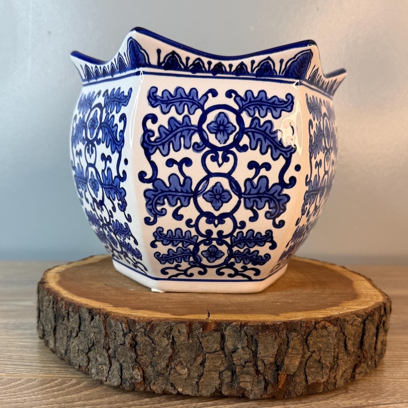 Puede incluir: Una maceta de cer&aacute;mica blanca y azul con borde festoneado. La maceta presenta un patr&oacute;n floral azul repetido. La maceta est&aacute; sobre una rodaja de madera redonda.