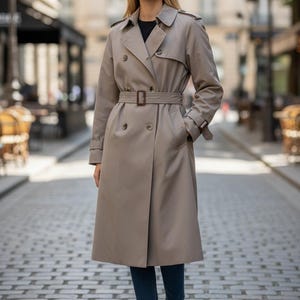 May include: A tan trench coat with a double-breasted design, a belt, and a classic collar. The coat is worn over a black top and blue jeans. White sneakers complete the look. The background is a cobblestone street.