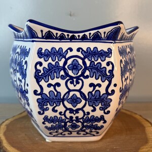 Puede incluir: Una maceta de cer&aacute;mica blanca con un dise&ntilde;o floral azul. La maceta tiene un borde festoneado &uacute;nico y una forma hexagonal. El patr&oacute;n azul presenta hojas y flores estilizadas. La maceta est&aacute; sobre una superficie de madera.