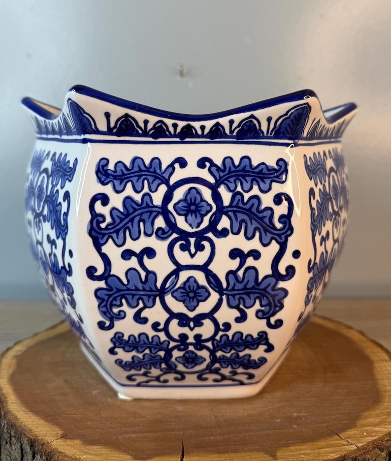 Puede incluir: Una maceta de cer&aacute;mica blanca con un dise&ntilde;o floral azul. La maceta tiene un borde festoneado &uacute;nico y una forma hexagonal. El patr&oacute;n azul presenta hojas y flores estilizadas. La maceta est&aacute; sobre una superficie de madera.