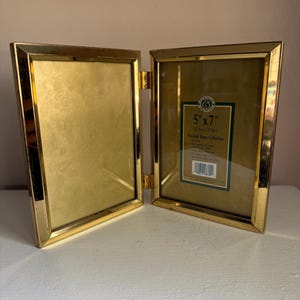 May include: A hinged, gold-colored metal photo frame, open to display two 5" x 7" (12.7cm x 17.8cm) pictures. The frame is from the "Polished Brass Collection" and has a label with the dimensions and a logo.