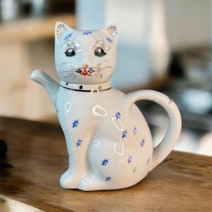 May include: A white ceramic cat-shaped teapot with blue floral accents. The teapot has a black collar with a gold clasp and features a detailed cat face with silver accents and green eyes. The teapot is sitting on a wooden surface.