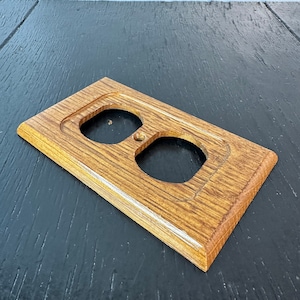 May include: A wooden electrical outlet cover with a light brown finish and a white border. The cover has two round openings for electrical outlets.