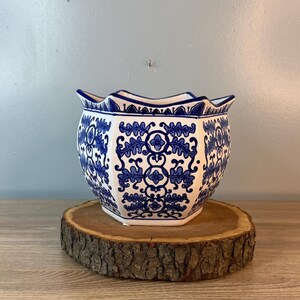 Puede incluir: Una maceta de cer&aacute;mica blanca y azul con borde festoneado. La maceta presenta un detallado patr&oacute;n floral azul. Se coloca sobre una rodaja de madera redonda y natural, mostrando su dise&ntilde;o decorativo. La maceta tiene forma hexagonal.