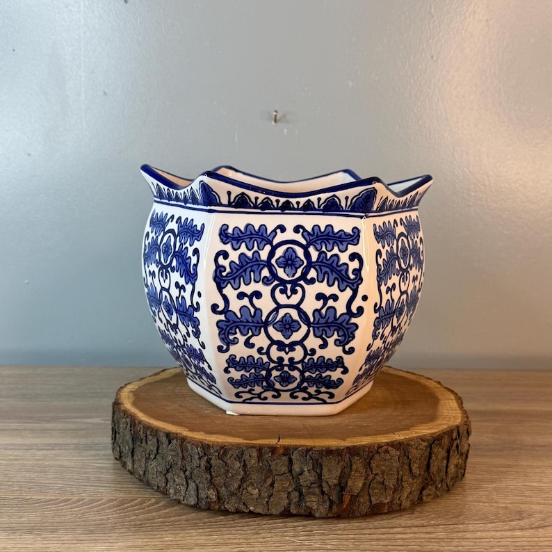 Puede incluir: Una maceta de cer&aacute;mica blanca y azul con borde festoneado. La maceta presenta un detallado patr&oacute;n floral azul. Se coloca sobre una rodaja de madera redonda y natural, mostrando su dise&ntilde;o decorativo. La maceta tiene forma hexagonal.