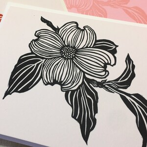May include: Four blank greeting cards with a black and white line drawing of a dogwood flower on the front. The cards are stacked on top of each other. The top card is white, the second card is pink, the third card is green, and the bottom card is red.