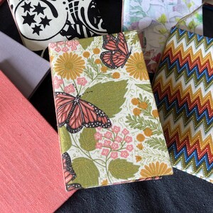 May include: A collection of rectangular notebooks featuring various covers, including solid coral, lavender, red, and blue, alongside patterned designs with butterflies, flowers, geometric zigzags, and black and white celestial motifs.