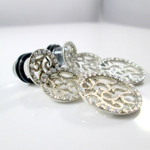 May include: Silver earrings with a filigree design and clear rhinestones. The earrings are dangling and have a circular shape.