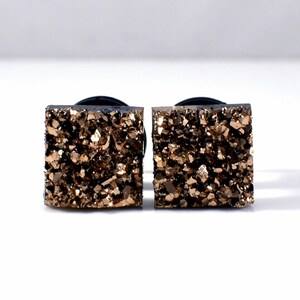 May include: A pair of square-shaped gold-toned earrings with a rough, textured surface. The earrings are made of a natural stone, possibly agate or quartz.