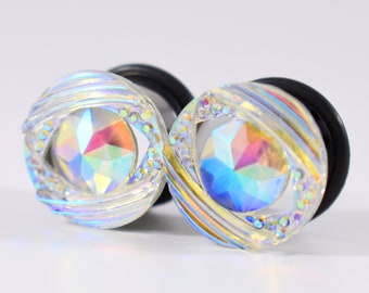 Puede incluir: Un par de dilatadores de oreja transparentes iridiscentes con un diseño de cristal. Los dilatadores tienen un respaldo de goma negra.