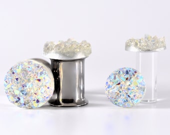 Puede incluir: Tres pares de dilatadores de oreja de cristal iridiscente. Los dilatadores están hechos de acero inoxidable y tienen un respaldo de acrílico transparente. El cristal es de color azul claro y blanco.