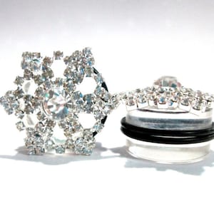 May include: A pair of silver ear gauges featuring a snowflake design. The snowflake is embellished with clear rhinestones. The gauges are made of acrylic and have a black rubber o-ring.