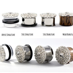 May include: Four different types of ear gauges with a crystal design. The gauges are made of acrylic, steel, wood, and steel. The acrylic gauge is a single flare, the steel gauges are single and double flare, and the wood gauge is a double flare.