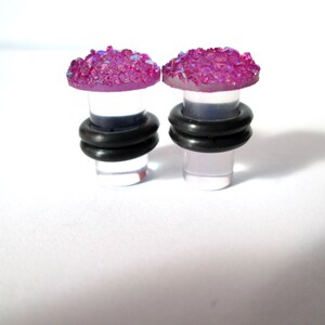 May include: Pair of clear acrylic ear gauges with a pink glitter top. The gauges have a black rubber o-ring around the base.