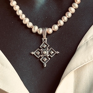 May include: A pearl necklace with a silver pendant. The pendant is a cross shape with a central gem and diamond-shaped accents. The necklace is composed of light pink and white pearls.