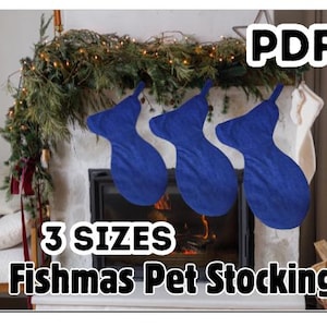 Pode incluir: Três meias azuis para animais de estimação Fishmas penduradas acima de uma lareira decorada com vegetação e luzes. A imagem inclui o texto "PDF", "3 TAMANHOS" e "Fishmas Pet Stocking".