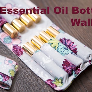 May include: Essential Oil Bottle Wallet with a floral pattern. The wallet is gray with a colorful floral design and holds several gold-capped essential oil bottles. The text "Essential Oil Bottle Wallet" is displayed in maroon.