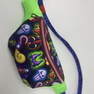 May include: A green and purple fanny pack with a zipper closure and a blue adjustable strap. The fanny pack has a black background with a colorful pattern of various Disney villains' icons.