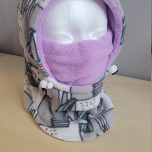 May include: A gray and white fleece hooded neck warmer with a purple fleece lining. The neck warmer has a unicorn print and two white toggles.