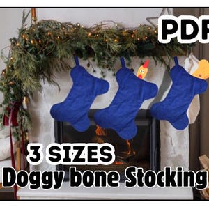 Peut inclure: Trois bas bleus en forme d'os de chien sont suspendus au-dessus d'une cheminée décorée de verdure et de lumières. Un bas a une décoration de poulet et un autre a un os. Le texte "PDF", "3 SIZES" et "Doggy bone Stocking" est affiché.