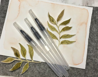 Op de afbeelding: Vier transparante aquarel penseelpennen met zwarte punten. De pennen zijn in een rij op een wit oppervlak gerangschikt met een aquarel achtergrond en groene bladeren ontwerpen.