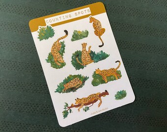 Puede incluir: Una hoja de pegatinas con siete leopardos ilustrados en varias poses, rodeados de follaje verde. Las pegatinas se titulan "Counting Spots" y son de "Julia K Art Studio".