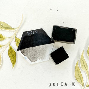 Puede incluir: Tres pastillas de pintura acuarela negra, dos cuadradas y una hexagonal, con la palabra "STEN" escrita en la pastilla hexagonal.
