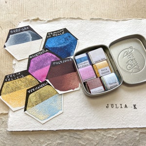 Puede incluir: Una colección de pinturas de acuarela en una pequeña lata de metal, con muestras de color individuales dispuestas alrededor. Las muestras son hexagonales y están etiquetadas con nombres como "Skele-Gro" y "Felix Felicis". Los colores incluyen azules, púrpuras, dorados y marrones.
