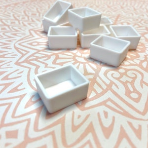 Pode incluir: Um conjunto de cubetas de tinta de aquarela de cerâmica branca. As cubetas estão dispostas em semicírculo sobre uma superfície com um padrão branco e rosa.