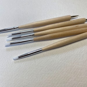 Op de afbeelding: Set van zes gereedschappen met houten handvatten en witte siliconen tips. De gereedschappen hebben verschillende vormen, waaronder een puntige tip, een ronde tip en een platte tip.