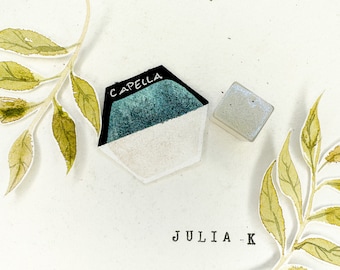 Può includere: Due pastiglie di acquerello, una con l'etichetta "CAPPELLA" in lettere nere. La pastiglia a sinistra è di colore blu-verde, e la pastiglia a destra è di colore bianco con una finitura scintillante. Le pastiglie sono su una superficie bianca con due foglie di acquerello verdi su entrambi i lati.
