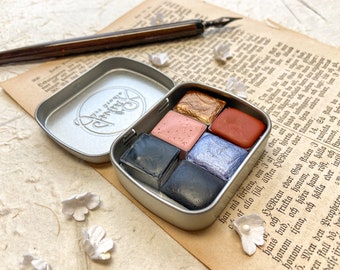 May include: A silver metal tin with a round logo that says "The Strange" in a cursive font. The tin contains six watercolor paint pans in a variety of colors, including gold, pink, red, black, and blue.