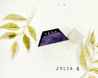 Può includere: Un panetto di acquerello viola e bianco con la parola "VEGA" stampata sulla parte superiore. Il panetto è su una superficie bianca con un piccolo panetto quadrato bianco accanto. Il testo "JULIA.K" è stampato sotto i panetti.