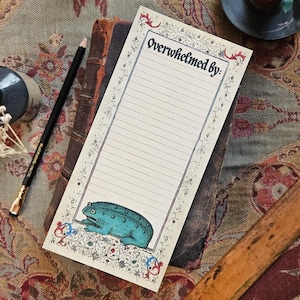 May include: A notepad featuring the text "Overwhelmed by:" and a teal frog illustration. The notepad has a decorative border and is placed on books. A pencil and other decorative items are also present, creating a vintage aesthetic.