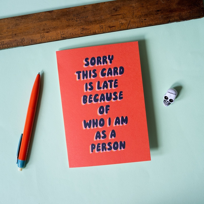 May include: Red card with the text "SORRY THIS CARD IS LATE BECAUSE OF WHO I AM AS A PERSON" in a black and pink font. A small white skull figurine is on a light blue surface.