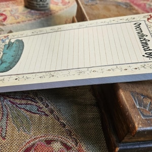 May include: A rectangular notepad with a cream background and lined pages. It features a teal pickle illustration and the words "Overwhelmed by:" in black script. The notepad has a decorative floral border.