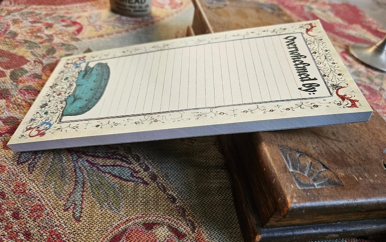 May include: A rectangular notepad with a cream background and lined pages. It features a teal pickle illustration and the words "Overwhelmed by:" in black script. The notepad has a decorative floral border.