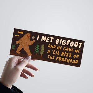 May include: A rectangular sticker with a dark brown background featuring a brown Bigfoot character holding a small white flower. The text reads: I MET BIGFOOT AND HE GAVE ME A 'LIL KISS ON THE FOREHEAD. Green pine trees and orange clouds are visible.