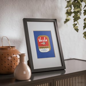 Puede incluir: Una impresi&oacute;n enmarcada de una etiqueta vintage de una lata de sardinas. La etiqueta es roja, amarilla y azul con el texto "Vesla Sardiner I Tomat" y un dibujo de un pez.