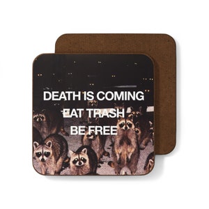 May include: A brown coaster featuring a black and white image of raccoons with glowing eyes. The text "DEATH IS COMING EAT TRASH BE FREE" is printed in white on the coaster.