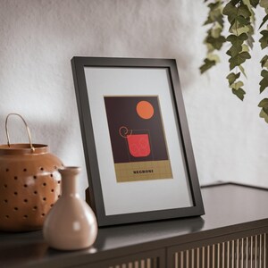 May include: A black framed print with a minimalist illustration of a cocktail glass with a red liquid and a yellow circle. The text "NEGRONI" is printed below the illustration.