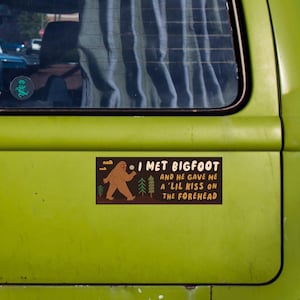 Puede incluir: Una pegatina rectangular con fondo marr&oacute;n oscuro que presenta una ilustraci&oacute;n de Bigfoot en color tostado, pinos verdes y el texto: I MET BIGFOOT AND HE GAVE ME A 'LIL KISS ON THE FOREHEAD. La pegatina est&aacute; sobre una superficie de metal verde lima.