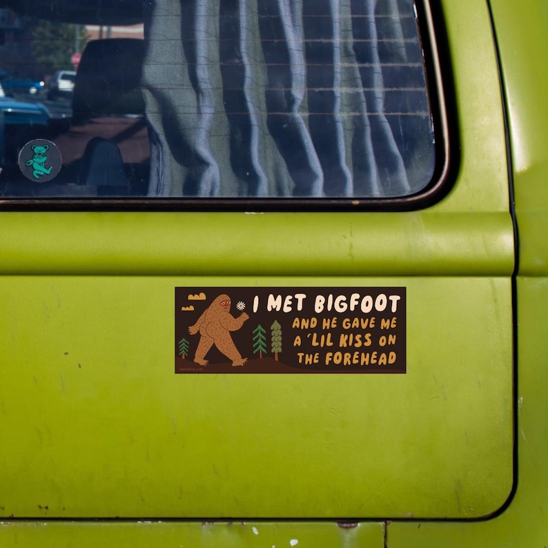 Puede incluir: Una pegatina rectangular con fondo marr&oacute;n oscuro que presenta una ilustraci&oacute;n de Bigfoot en color tostado, pinos verdes y el texto: I MET BIGFOOT AND HE GAVE ME A 'LIL KISS ON THE FOREHEAD. La pegatina est&aacute; sobre una superficie de metal verde lima.