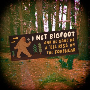 Puede incluir: Una pegatina rectangular con fondo marr&oacute;n oscuro que presenta una ilustraci&oacute;n de dibujos animados de un Bigfoot marr&oacute;n caminando junto a pinos verdes. El texto dice: I MET BIGFOOT AND HE GAVE ME A 'LIL KISS ON THE FOREHEAD en letras color crema.