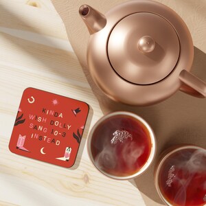 Pu&ograve; includere: Un sottobicchiere quadrato rosso con sfondo bianco e testo nero che recita "Kinda wish Dolly sang 10-3 instead". Il sottobicchiere ha un ferro di cavallo rosa, una stella rosa, un cappello da cowboy rosa e stivali da cowboy rosa. Il sottobicchiere &egrave; appoggiato su una superficie di legno accanto a una teiera di rame e due tazze di t&egrave;.