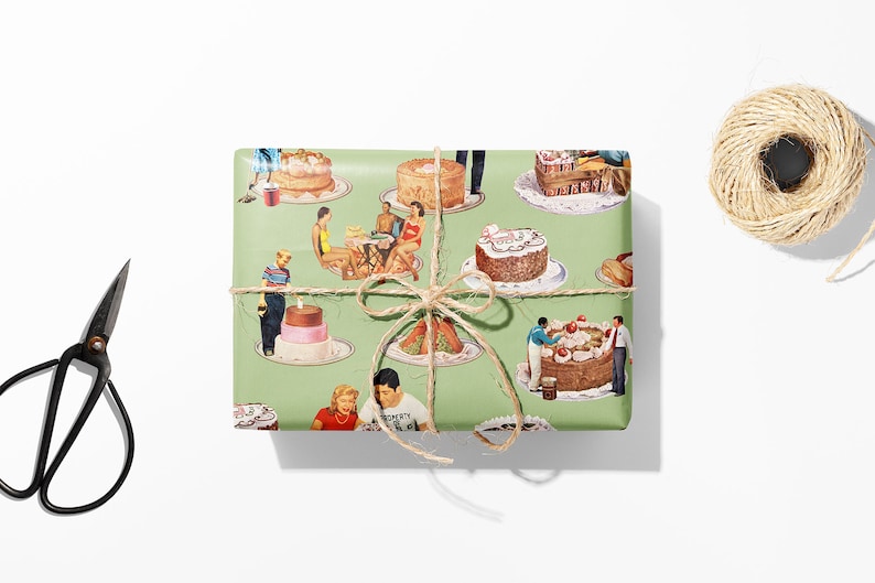 May include: A wrapped gift with a green background featuring a vintage cake design. The gift is tied with twine and a bow. Black scissors and a spool of twine are on a white surface.