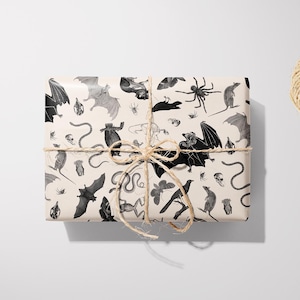 May include: A wrapped gift box with a beige background, featuring a repeating pattern of bats, spiders, snakes, and other Halloween-themed illustrations in black and gray. The box is tied with twine, and a pair of black scissors and a ball of twine are nearby.