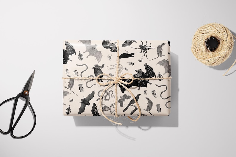 May include: A wrapped gift box with a beige background, featuring a repeating pattern of bats, spiders, snakes, and other Halloween-themed illustrations in black and gray. The box is tied with twine, and a pair of black scissors and a ball of twine are nearby.